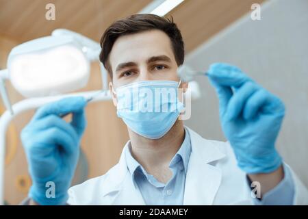 Dentista maschile professionale in camice bianco, maschera protettiva e guanti tenere strumenti dentali e iniziare il lavoro Foto Stock
