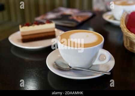 XtWhite tazza di caffè e pezzo di torta a strisce sul tavolo in caffetteria su sfondo sfocato. Messa a fuoco superficiale. Foto Stock