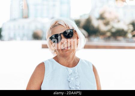 Ritratto di bionda positivo soddisfatta donna matura in occhiali da sole sguardi alla telecamera all'aperto nel parco Foto Stock