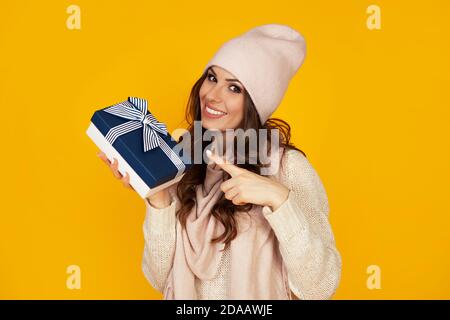 Felice giovane donna sorridente puntando dito tenendo in mano la scatola regalo blu. Un dono al tuo amato uomo. Una ragazza in un maglione guarda la fotocamera e punta a un regalo. Natale Capodanno concetto di vacanza Foto Stock