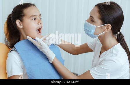 Millennial signora medico in uniforme, maschera protettiva e guanti di gomma esamina di bambino piccolo con bocca aperta Foto Stock