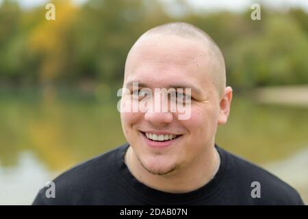 Felice giovane uomo casual con testa rasata in piedi all'aperto un parco vicino a un lago ridendo Foto Stock