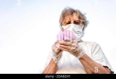 La vecchia donna in maschera e guanti tiene la palla assomiglia al coronavirus nelle sue mani. Le persone anziane rischiano di infettarsi quando lasciano casa Foto Stock
