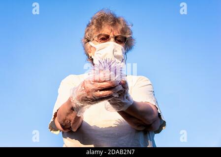 Protetto da una maschera e guanti, la vecchia donna tiene una palla assomiglia al coronavirus nelle sue mani. Foto Stock