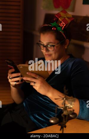 Donna giovane sorridente con capelli decorati che guarda in due telefoni cellulari a natale. Fuoco su primo piano Foto Stock