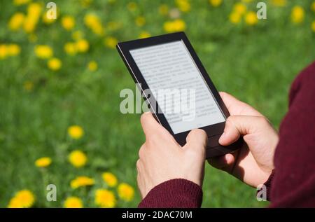 Un uomo sta leggendo un libro elettronico in primavera giorno di sole. Testo sfocato. Solo le sue mani sono sulla foto. Campo dente di leone sullo sfondo. Foto Stock