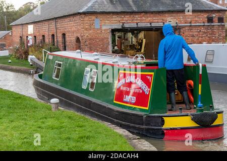 una barca stretta sul canale grand union con un uomo che indossa una giacca impermeabile che guida la barca in un blocco a braunston. Foto Stock