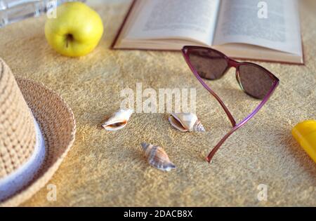 Cappello di paglia, conchiglie, crema solare, occhiali da sole, mela, bottiglia d'acqua e libro aperto su asciugamano beige. Luce solare soffusa. Foto Stock