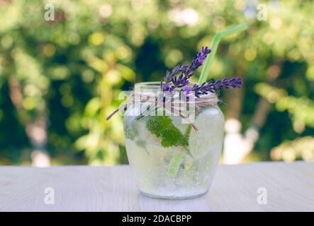 Vaso di vetro con fresca limonata fatta in casa di menta, balsamo di limone, limone, lime, decorato con fiori di lavanda su tavolo di legno nel giardino Foto Stock
