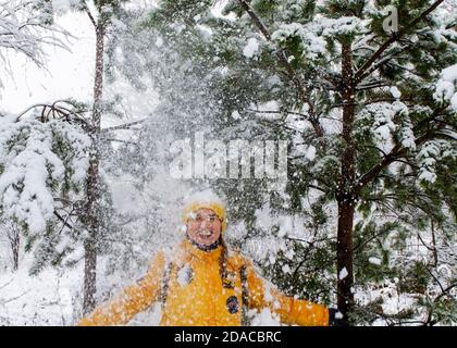 Giovane donna caucasica sorridente felice in giacca gialla sotto la neve caduta da rami di pino nella foresta invernale. Emozioni positive, camminando in un'erba innevata Foto Stock