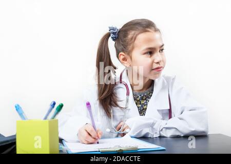 Prescolare ragazza età giocare carattere doctror, camice bianco laboratorio, seduta alla scrivania, uno sfondo bianco Foto Stock