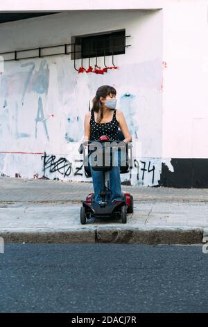 Giovane donna in scooter elettrico per disabili. Non può attraversare la strada perché non c'è rampa. Grunge parete sullo sfondo. Foto Stock