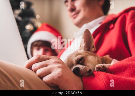 Cane cucciolo che dorme con l'uomo e il bambino il giorno di Natale al computer portatile, seduto su un divano nel soggiorno con l'albero di Natale a casa. Foto Stock