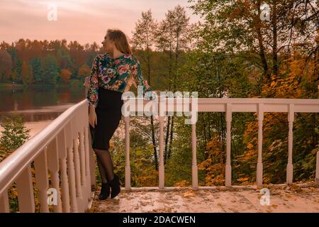 Una giovane donna allegra con un aspetto carismatico in un la camicia colorata si appoggia contro una ringhiera bianca appoggiata a una ringhiera bianca contro Foto Stock