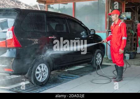 Il pulitore per auto con l'uniforme rossa spruzzi alta pressione l'acqua, utilizzando un tubo flessibile, alla carrozzeria della vettura nel autolavaggio Foto Stock