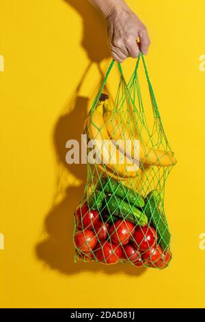 Mano maschile che tiene una borsa bianca in rete con verdure su sfondo giallo con spazio per la copia. Concetto di shopping senza sprechi. Foto Stock