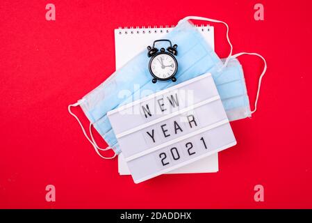Scatola luminosa con l'iscrizione Capodanno, maschera facciale da virus, orologio su sfondo rosso. Consapevolezza del nuovo anno 2021. Coronavirus pandemico nel 2021. C Foto Stock