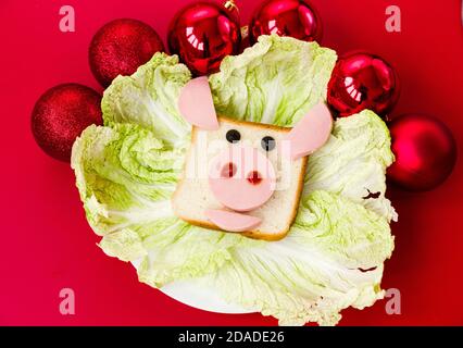 Un sandwich a forma di testa di un toro su un piatto decorato con calchi di insalata. La piastra si trova su uno sfondo rosso. Palline di natale rosse decorano il Foto Stock