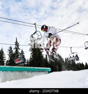 Sciatore maschile in giacca da sci e casco facendo saltare il giorno d'inverno. Uomo freerider saltando in aria presso la stazione sciistica con pini e impianti di risalita sullo sfondo. Concetto di sport invernale estremo Foto Stock