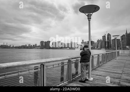 NEW YORK, USA - 27 aprile 2016: Un fotografo maschile anziano si erge su un molo e scatta una foto della vista panoramica dello skyline di Midtown Manhattan. Vista f Foto Stock