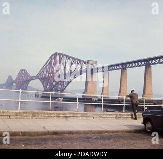 Anni '60, storico, un uomo in piedi accanto alle ringhiere che si affaccia sulle acque del Firth of Forth e su al Forth Bridge, un ponte ferroviario a sbalzo attraverso l'estuario, vicino a Edimburgo, Scozia, Regno Unito. Quando fu inaugurato nel 1890, aveva la più lunga campata di ponte a sbalzo al mondo. Foto Stock
