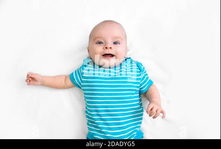 Un bambino positivo che indossa una t-shirt a righe blu giace su un letto bianco, sorridente e guardando la macchina fotografica. Felice infanzia. Vista dall'alto Foto Stock