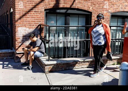 NEW YORK, USA - 22 settembre 2017: Scena di strada a Dumbo, Brooklyn. Un giovane con un telefono cellulare si siede sul parapetto di una casa in mattoni e una donna nera Foto Stock