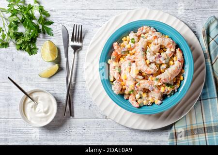 Insalata di gamberi con salsa di panna acida e succo di lime, mais, cipolla rossa e prezzemolo serviti su un recipiente per insalata blu su un piatto di ricarica bianco con posate o Foto Stock