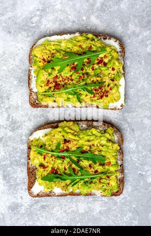 Sandwich a colazione - fetta di pane scuro a grani interi, formaggio cremoso, guakomole, guarnito con rucola, paprika su tavolo in cemento Vista dall'alto Foto Stock