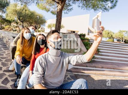 Gruppo di giovani amici caucasici seduti su una piazza della città parete che indossa covid-19 coronavirus maschere faccia facendo auto ritratto con smartphone per un'immagine nitida Foto Stock