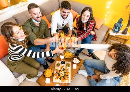 Gruppo di giovani che tostano per aperitivo a casa - Mixed race amici bere cocktail spritz seduta sul divano con Spuntini che indossano il Covid-1 abbassato Foto Stock