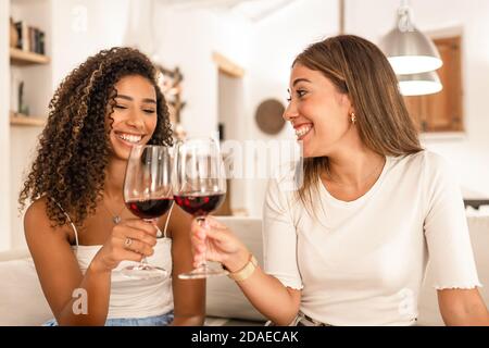 Due giovani donne di razza mista che festeggiano a casa tostando con Bicchieri da vino rosso - bruna cagliata ispanica sorridente il suo migliore amico seduto Foto Stock