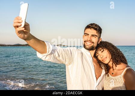 Misto corsa coppia in vacanza al mare resort facendo un Autoritratto al tramonto o all'alba - bel sorriso bearded Uomo scatta foto con il suo bebaut ispanico Foto Stock