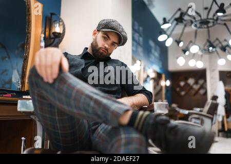 Il barbiere con abiti eleganti si siede sulla sedia nel salone Foto Stock