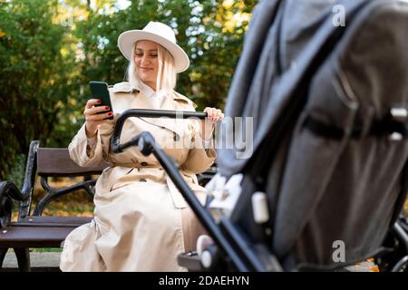 La madre felice con uno smartphone nelle mani siede su un panca nel parco e scogli il bambino in un passeggino Foto Stock