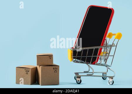 Carrello giocattolo con scatole e smartphone con schermo vuoto su sfondo blu. Spazio di copia per testo o disegno. Vendita, sconto, acquisto e consegna Foto Stock