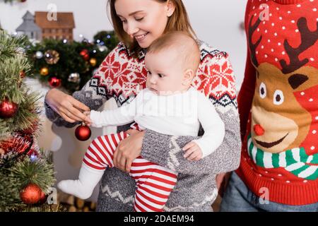 donna felice che tiene in armi bambino ragazzo mentre decorando natale albero vicino a marito Foto Stock