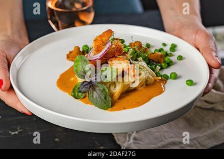 Le mani delle donne tengono un piatto di pasta e gamberi fritti con piselli verdi e salsa originale di chanterelles su uno sfondo scuro del tavolo. Foto Stock