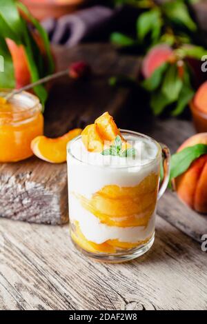 Dessert alla frutta di pesca in una tazza di vetro su un tavolo rustico in legno con frutta fresca di pesca, marmellata di pesche. Dessert fatto in casa con frutta. Macedonia di frutta con yogurt o. Foto Stock