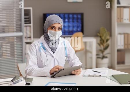 Ritratto di medico musulmano in maschera protettiva seduto al tavolo con tablet digitale mentre si lavora in ospedale Foto Stock