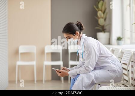 Medico femminile in camice bianco e in maschera digitando A. messaggio sul suo telefono cellulare seduto in corridoio Foto Stock