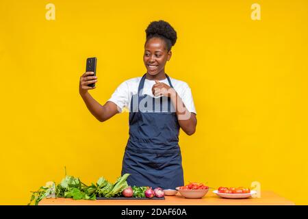 chef donna africana che fa una videochiamata con il suo telefono con verdure e altri prodotti alimentari di fronte a lei su una scrivania Foto Stock