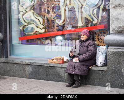 Kiev, Ucraina - 16 novembre 2019: Persone svantaggiate e impoverite sulla strada di Kiev. Una donna anziana si siede vicino a una vetrina e vende noci e. Foto Stock
