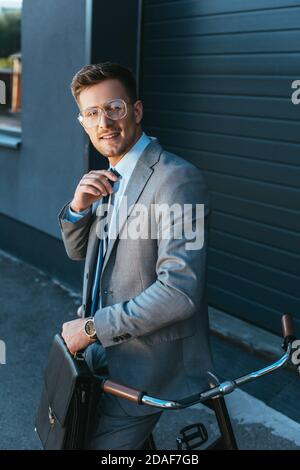 Uomo d'affari sorridente con borsa portadocumenti regolabile vicino alla bicicletta all'aperto Foto Stock