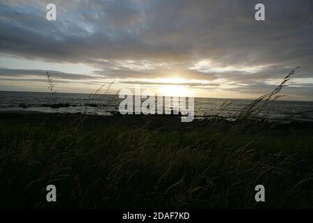 La costa dell'Ayrshire si affaccia su Ailsa Craig, Scozia, Regno Unito orizzonte ampio con erba in primo piano, Firth di Clyde a metà distanza e il tramonto del sole dietro Ailsa Craig Foto Stock