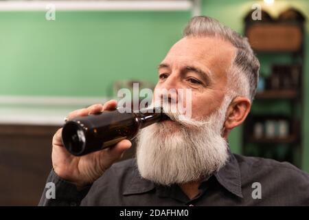Vecchio uomo caucasico bearded, modello bearded, in un bar retrò hipster beve una bottiglia di birra, ritratto fotografico. Foto Stock