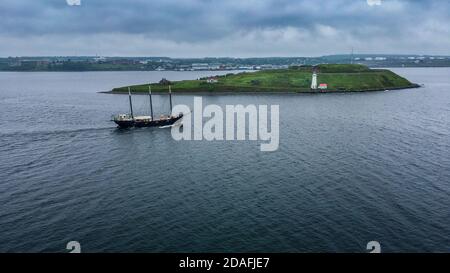 Halifax, Canada--18 giugno 2018; tre passi di barca a vela alta alberata di fronte a Georges Island con tetto rosso faro bianco che si dirige verso il mare Foto Stock