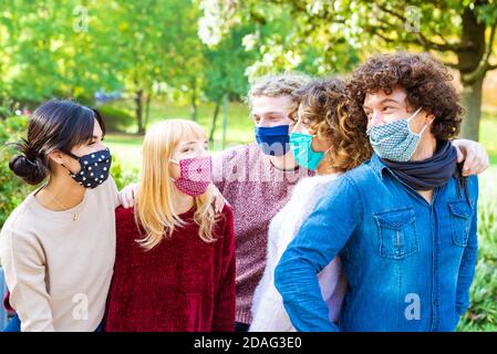 I giovani si divertono intorno al parco della città durante il coronavirus Outbreak.Happy amici multietnici indossare maschere di protezione del viso e ridere insieme. Foto Stock