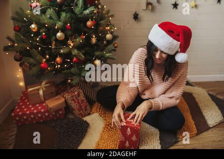 Felice giovane donna nel cappello di santa che avvolge il regalo di natale in carta moderna sotto l'albero di natale con le luci nella stanza decorata festa. Vacanze invernali pre Foto Stock