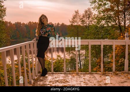 Felice donna si appoggia contro una ringhiera bianca giovane guardando fotocamera bionda in una camicia colorata, sullo sfondo del lago innocente autunno Foto Stock